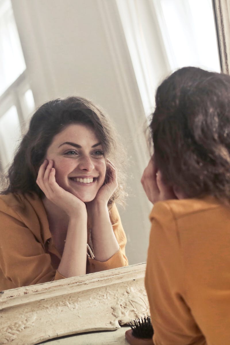 A cheerful woman smiles at her reflection in a vintage-style mirror, exuding positivity and warmth.